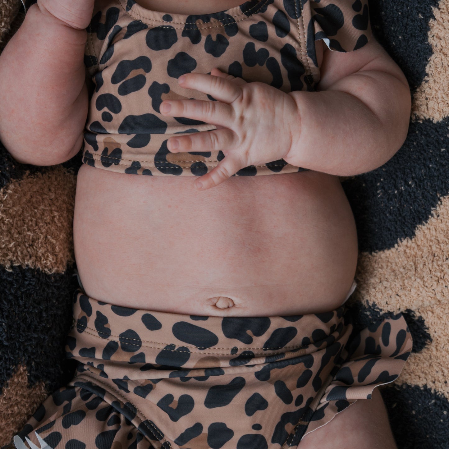 A close-up of a baby wearing the forever french baby *PRE-ORDER* Girl's Two Piece Swimsuit in leopard print, showing the baby's belly, hand, and arm while lying on a matching textured surface.