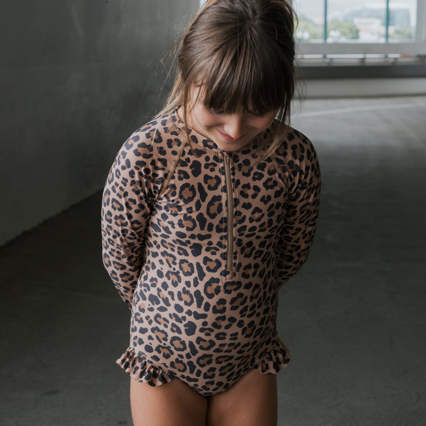A young girl with brown hair and bangs smiles indoors, wearing the forever french baby *PRE-ORDER* Girl's Long Sleeve Swimsuit | Leopard—an adorable one-piece with leopard print and ruffled hips, her hands tucked behind her back.