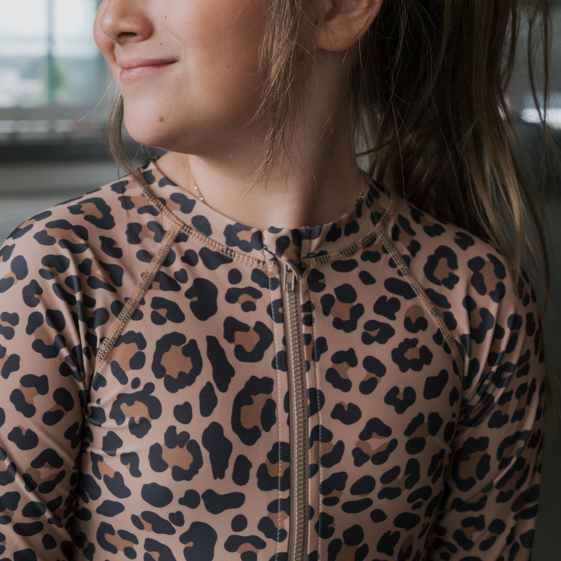 A young girl wears the forever french baby *PRE-ORDER* Girl's Long Sleeve Swimsuit | Leopard, smiling softly and looking to the side. Only her face and upper torso are visible.