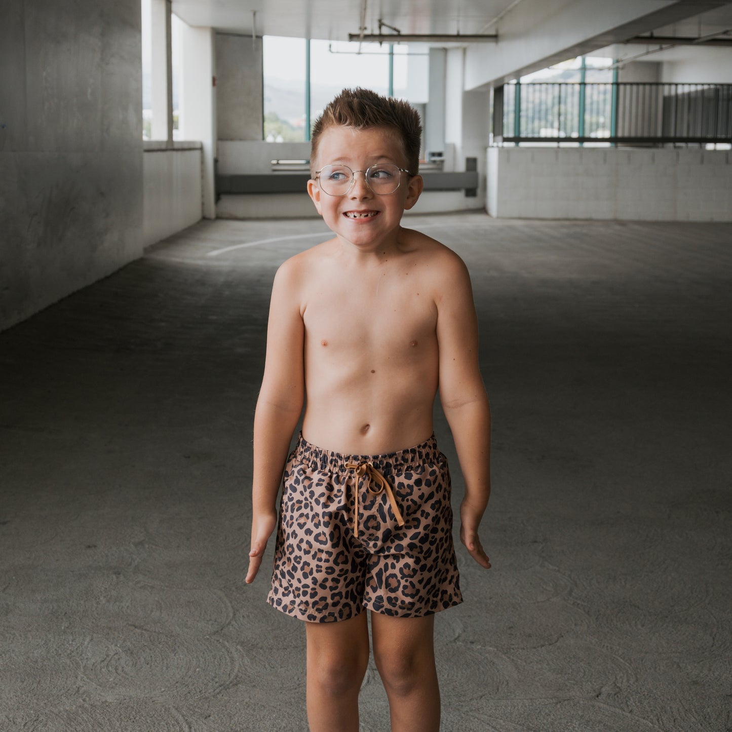 A young boy wears forever french baby’s *PRE-ORDER* Child Board Short | Leopard, standing indoors on concrete with glasses, arms at his sides and a wide, awkward grin.