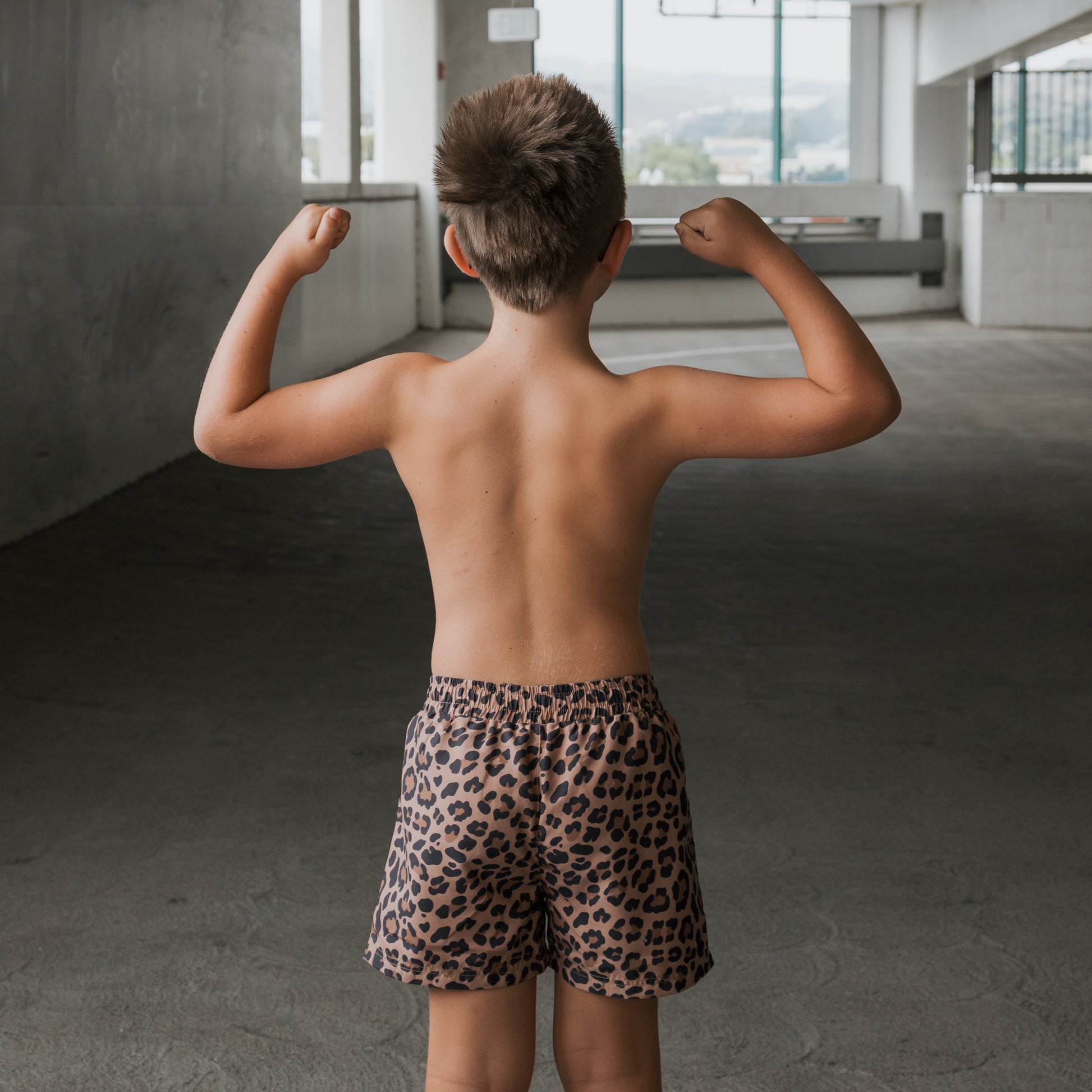 A child models the forever french baby *PRE-ORDER* Child Board Short | Leopard, standing in an open area facing away and flexing their arms to show off their muscles.
