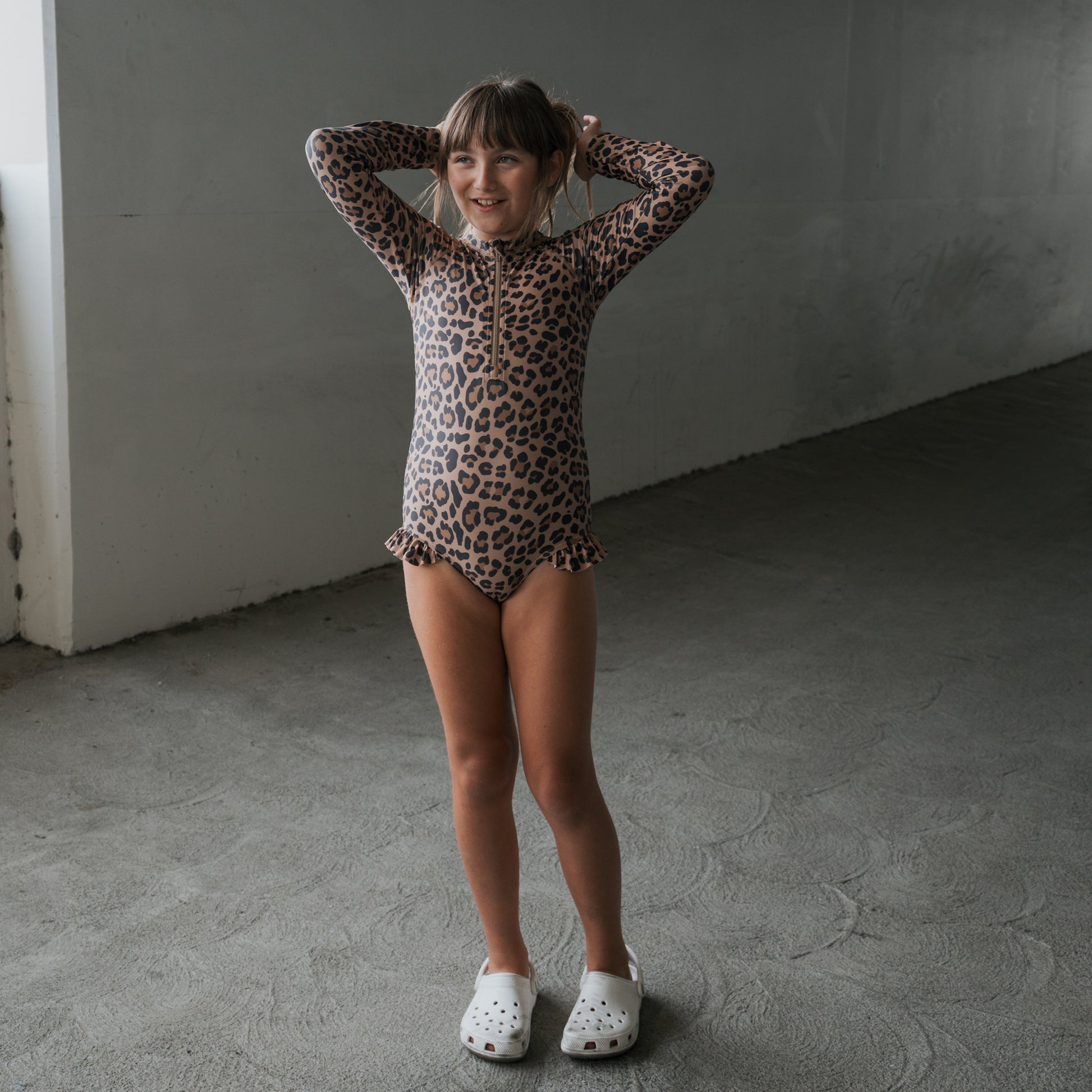 A young girl smiles indoors on a concrete floor, wearing the *PRE-ORDER* Girl's Long Sleeve Swimsuit | Leopard from forever french baby with white clogs, her hands resting behind her head.