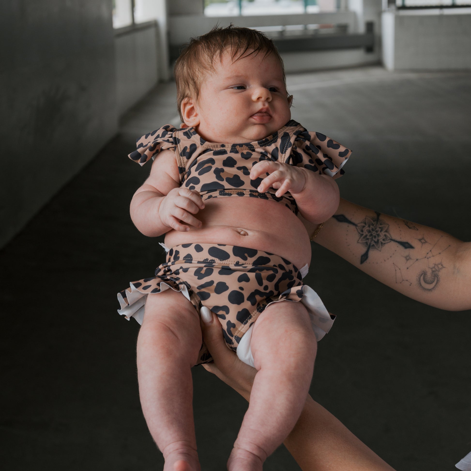 A baby in the *PRE-ORDER* Girl's Two Piece Swimsuit | Leopard by forever french baby is held up by a tattooed adult in a bright indoor setting.