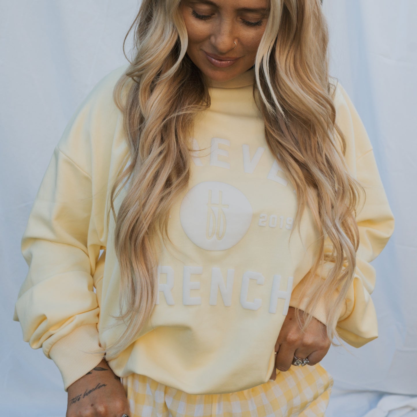 A person with long, wavy blonde hair wears the forever french baby Adult Sweatshirt in ff Yellow and matching yellow gingham pants, standing before a light fabric backdrop. The sweatshirt features white "2019 FRENCH" text.