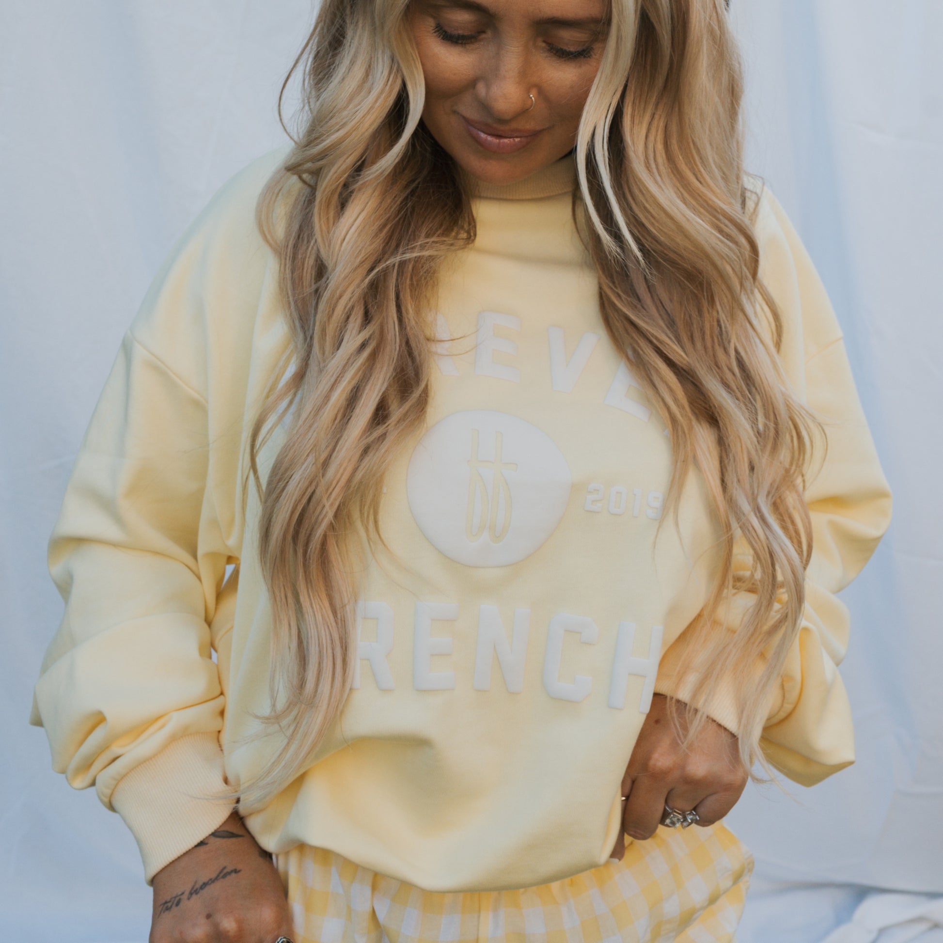 A person with long, wavy blonde hair wears the forever french baby Adult Sweatshirt in ff Yellow and matching yellow gingham pants, standing before a light fabric backdrop. The sweatshirt features white "2019 FRENCH" text.