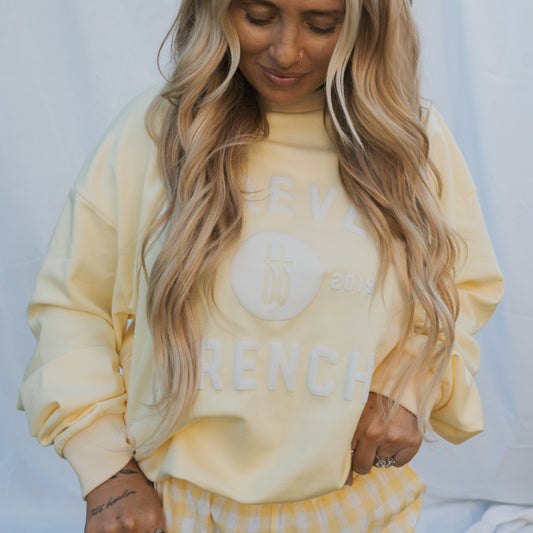A person with long, wavy blonde hair wears the forever french baby Adult Sweatshirt in ff Yellow and matching yellow gingham pants, standing before a light fabric backdrop. The sweatshirt features white "2019 FRENCH" text.