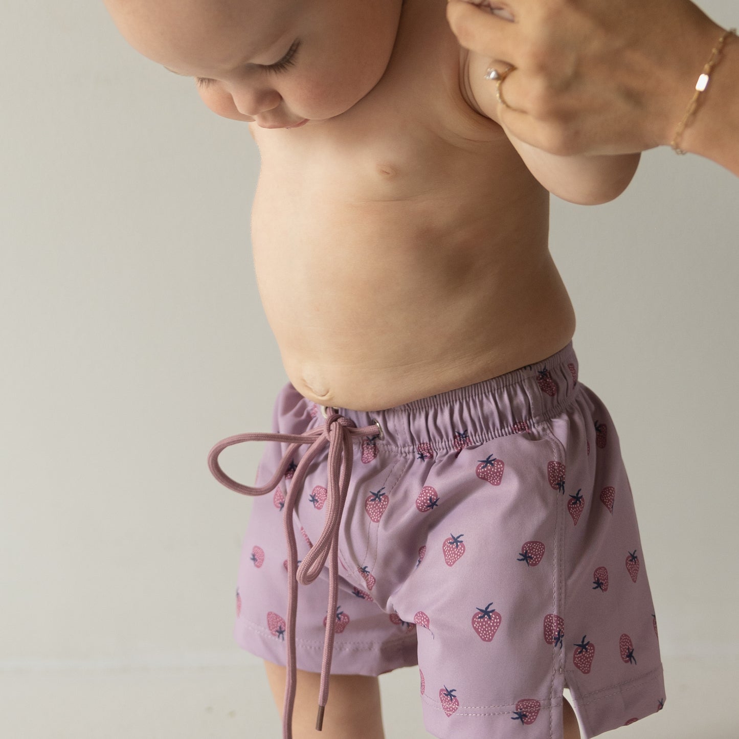 A baby wears forever french baby's Matilda Child Board Short—a light purple swim short with a strawberry print—gently held by an adult against a plain background, showcasing adorable kids’ summer swimwear.