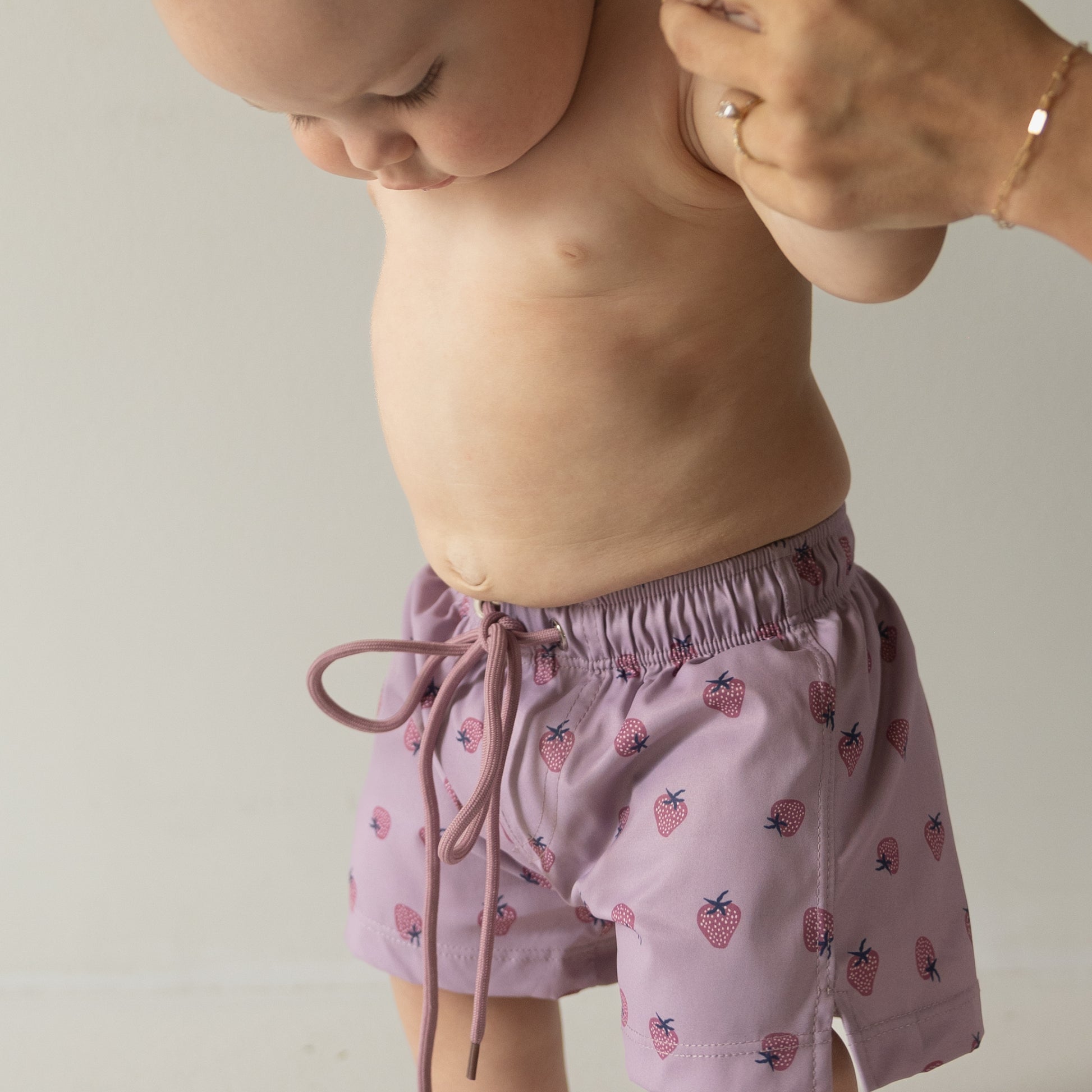 A baby wears forever french baby's Matilda Child Board Short—a light purple swim short with a strawberry print—gently held by an adult against a plain background, showcasing adorable kids’ summer swimwear.