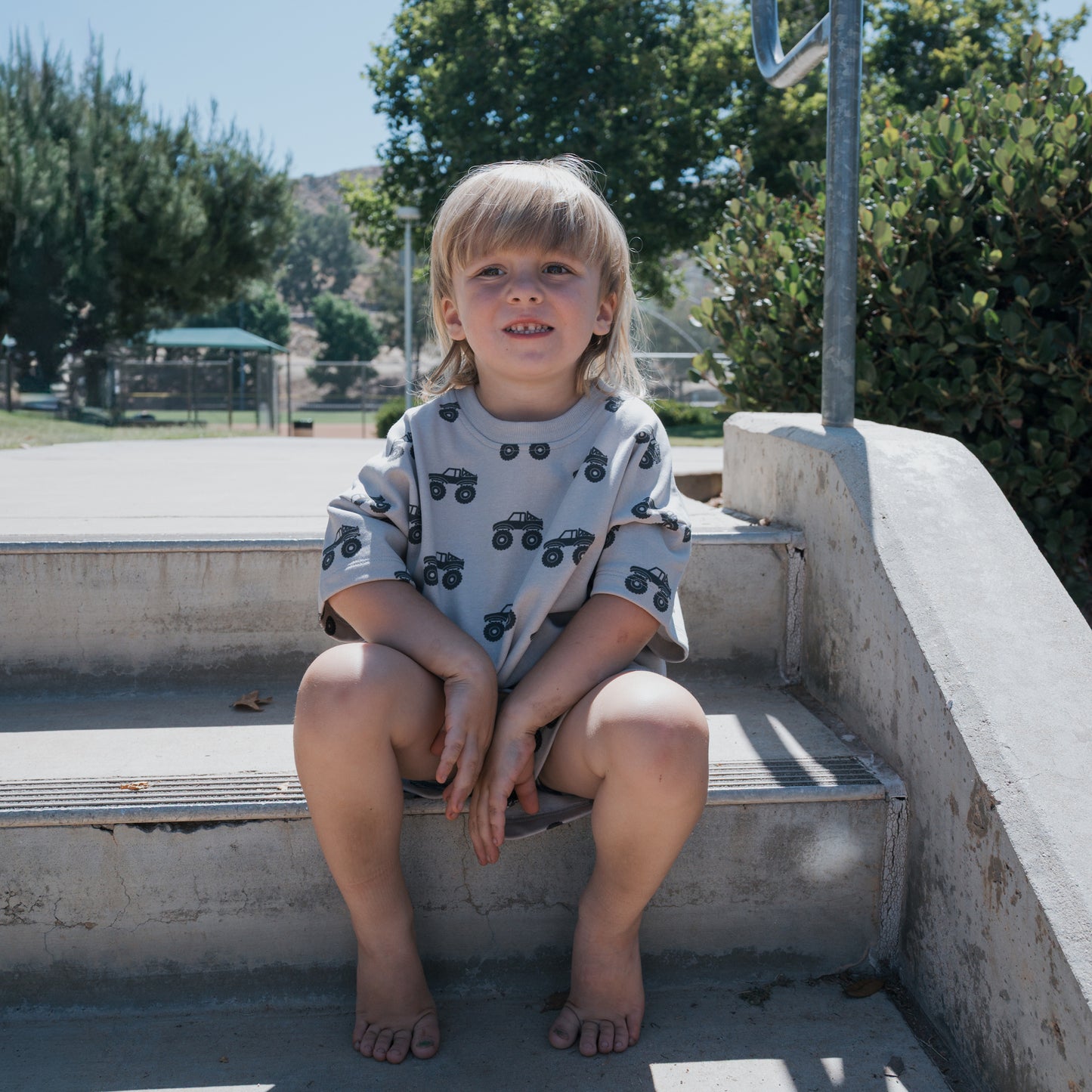 A young child with long blond hair, wearing the forever french baby French Terry Short Set | Monster Truck, sits barefoot on concrete steps outdoors, smiling. Trees and a sunny park are visible in the background.