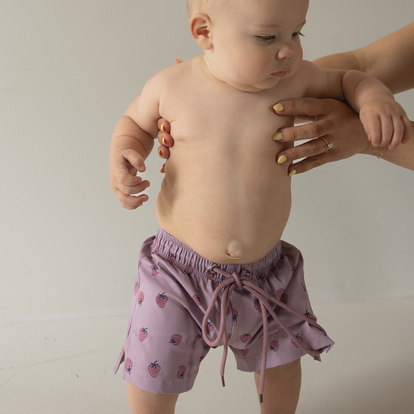 A baby wears the Matilda Child Board Short by forever french baby, featuring a fruit pattern in light purple, held upright by an adult with yellow-painted nails. A plain neutral background highlights this adorable summer look.