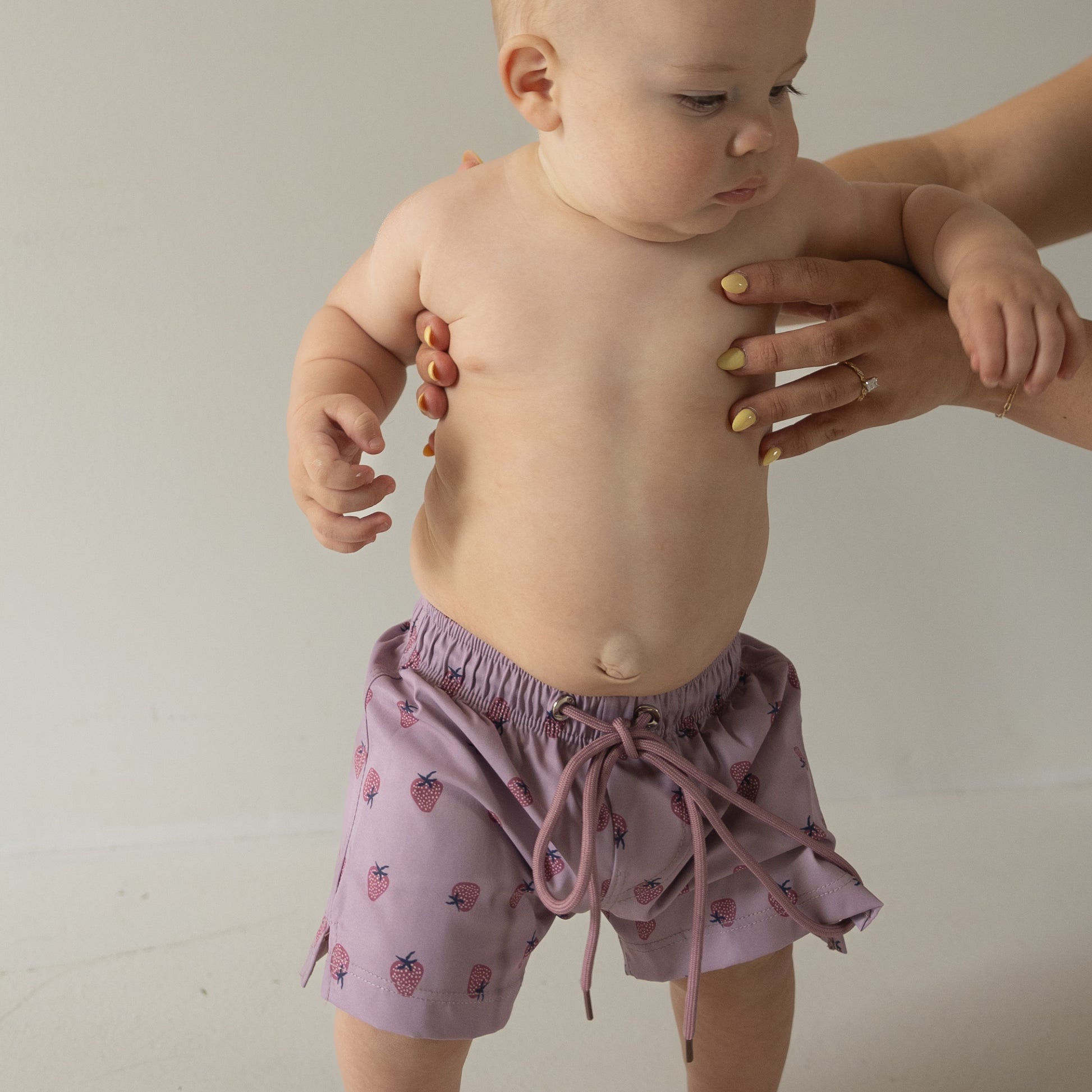 A baby wears the Matilda Child Board Short by forever french baby, featuring a fruit pattern in light purple, held upright by an adult with yellow-painted nails. A plain neutral background highlights this adorable summer look.
