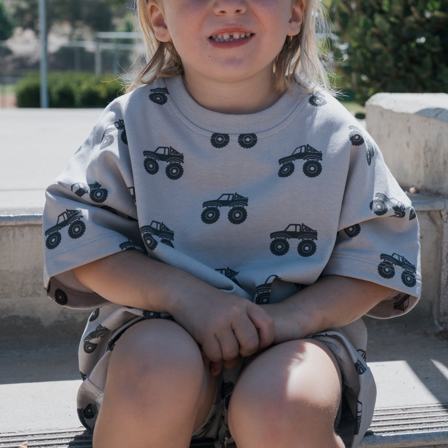 A young child with long blonde hair sits on outdoor steps, smiling slightly, wearing the forever french baby French Terry Short Set | Monster Truck—featuring a light gray short sleeve top and shorts with black monster truck prints.