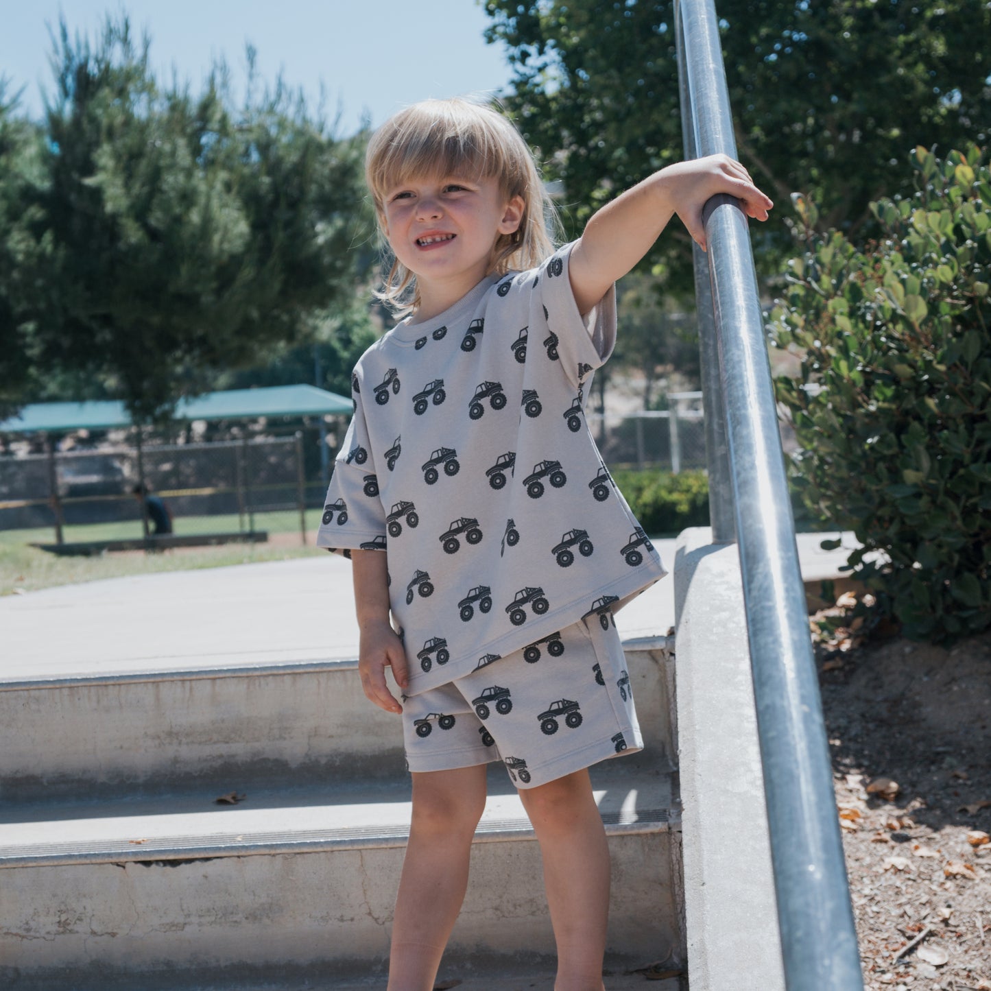 A young child with long blonde hair smiles while standing barefoot on outdoor steps, wearing the forever french baby French Terry Short Set | Monster Truck— a matching light gray top and shorts with black car prints. Trees and a park are in the background.