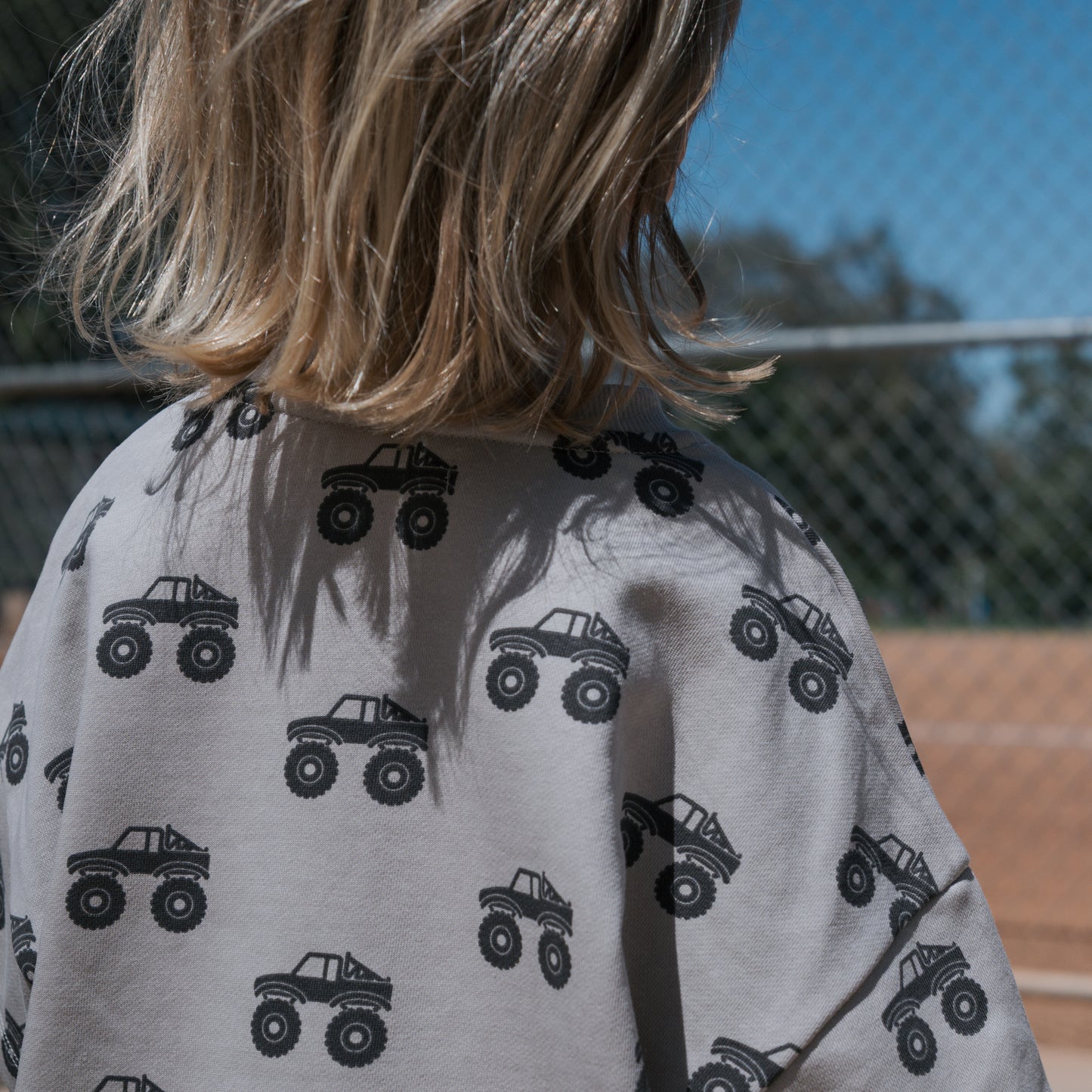 A blond-haired child, seen from behind, wears the forever french baby French Terry Short Set | Monster Truck—light gray short sleeves and shorts with black monster trucks—standing outdoors near a metal fence on a sunny day.