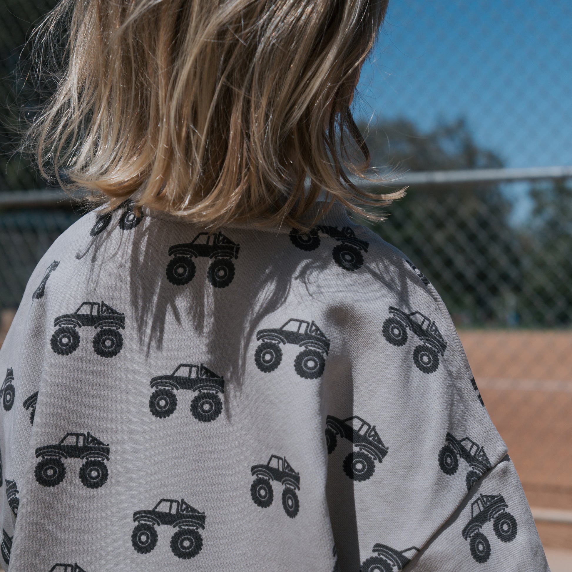 A blond-haired child, seen from behind, wears the forever french baby French Terry Short Set | Monster Truck—light gray short sleeves and shorts with black monster trucks—standing outdoors near a metal fence on a sunny day.