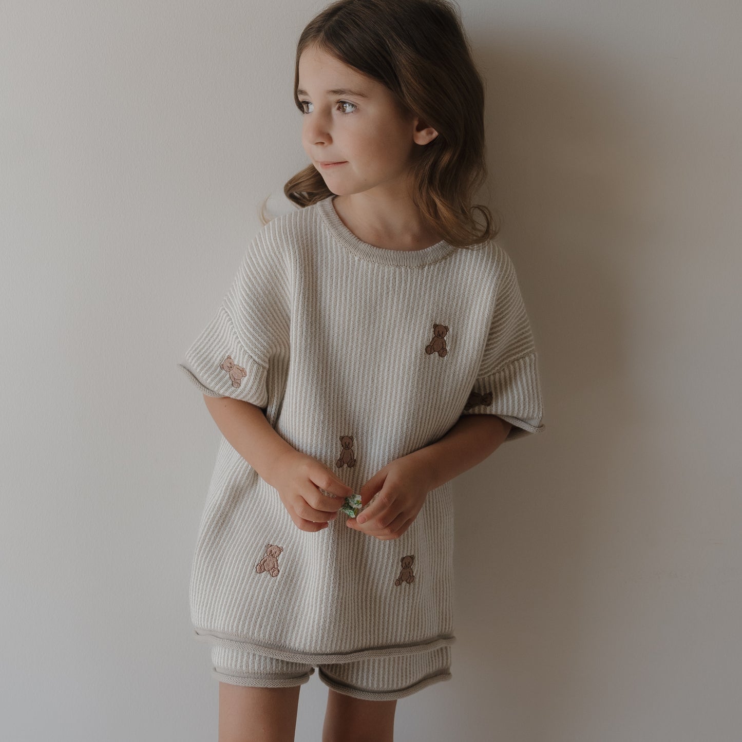A young girl with long brown hair stands against a plain wall, wearing the forever french baby Knit Short Set | Teddy, featuring teddy bear prints and drawstring shorts, while holding a small green object in her hands.