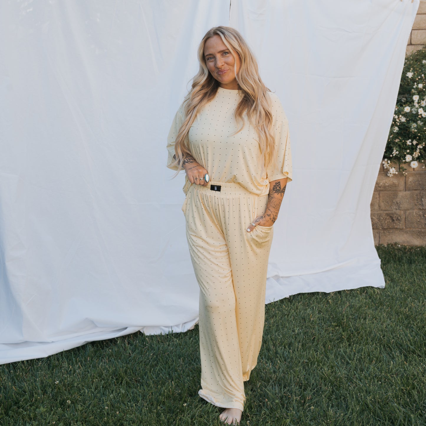 A woman with long blonde hair stands on grass before a white fabric backdrop, wearing the Women's Boxy Tee Lounge Set by forever french baby in light yellow with small dots. White flowers and a stone wall can be seen in the background.