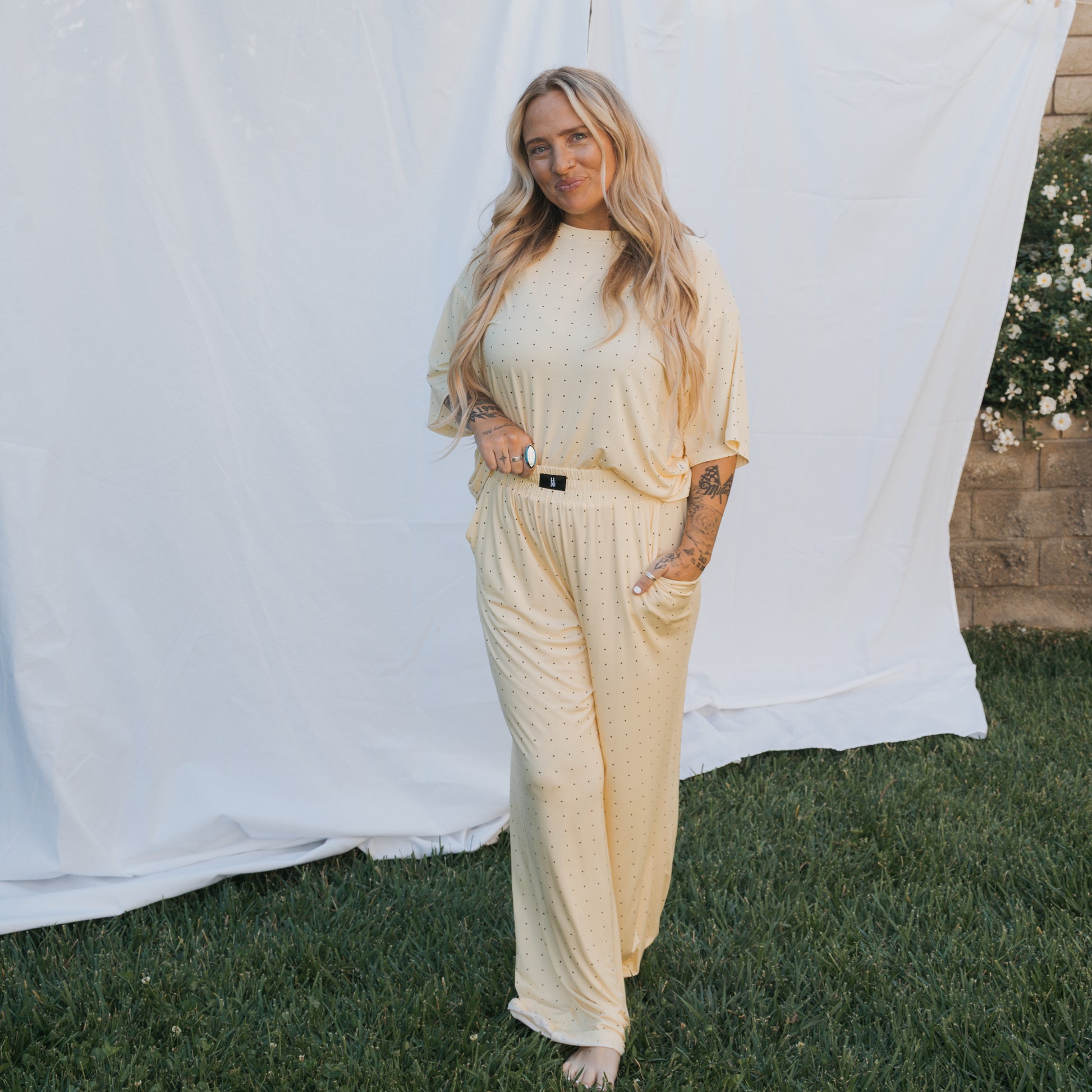 A woman with long blonde hair stands on grass before a white fabric backdrop, wearing the Women's Boxy Tee Lounge Set by forever french baby in light yellow with small dots. White flowers and a stone wall can be seen in the background.