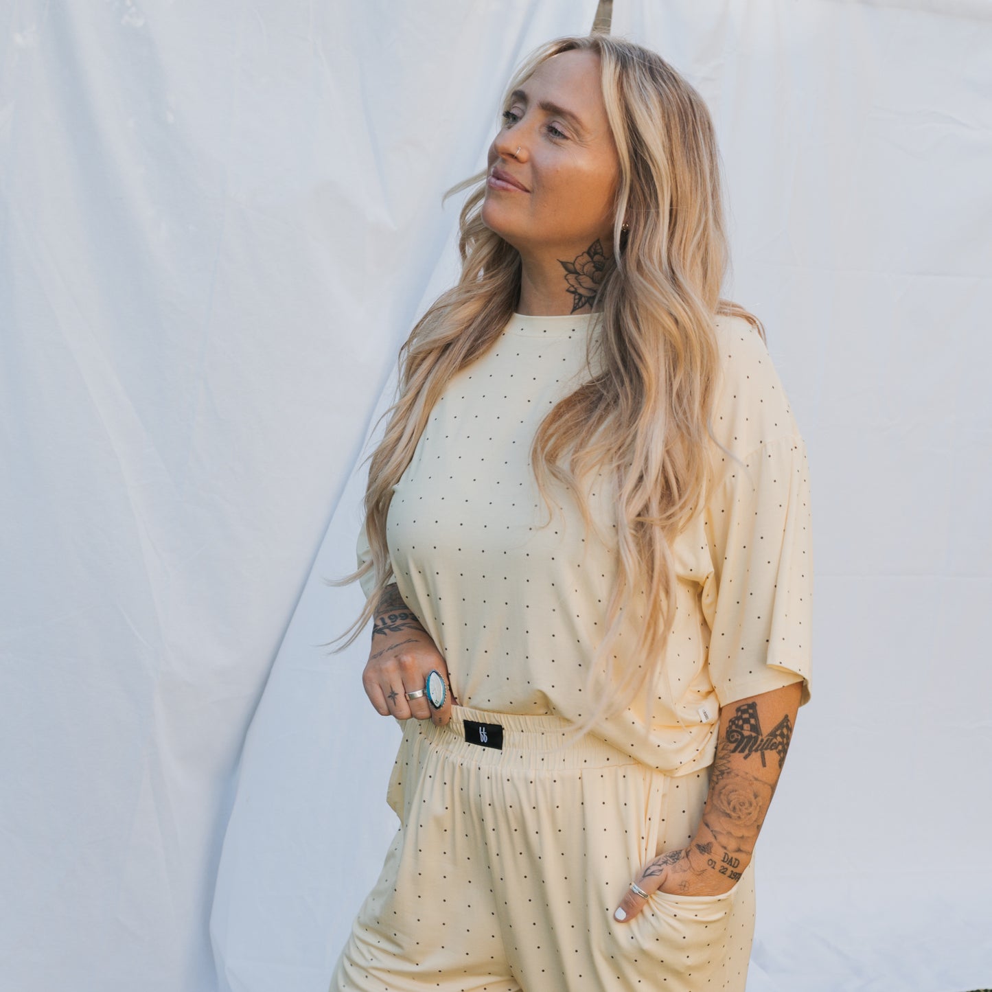 A person with long, wavy blonde hair and tattoos stands in front of a white backdrop, wearing the Women's Boxy Tee Lounge Set by forever french baby in a soft yellow polka dot print, radiating comfort and laid-back style.