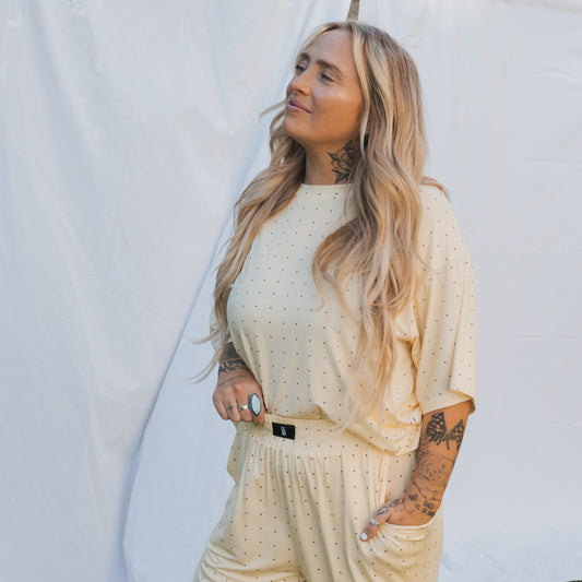 A person with long, wavy blonde hair and tattoos stands in front of a white backdrop, wearing the Women's Boxy Tee Lounge Set by forever french baby in a soft yellow polka dot print, radiating comfort and laid-back style.