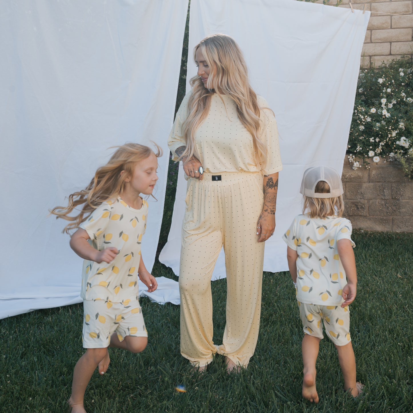 A woman in a Women's Boxy Tee Lounge Set by forever french baby stands on grass, watching two children in matching bamboo pajamas play with a white sheet and blooming flowers in the background.