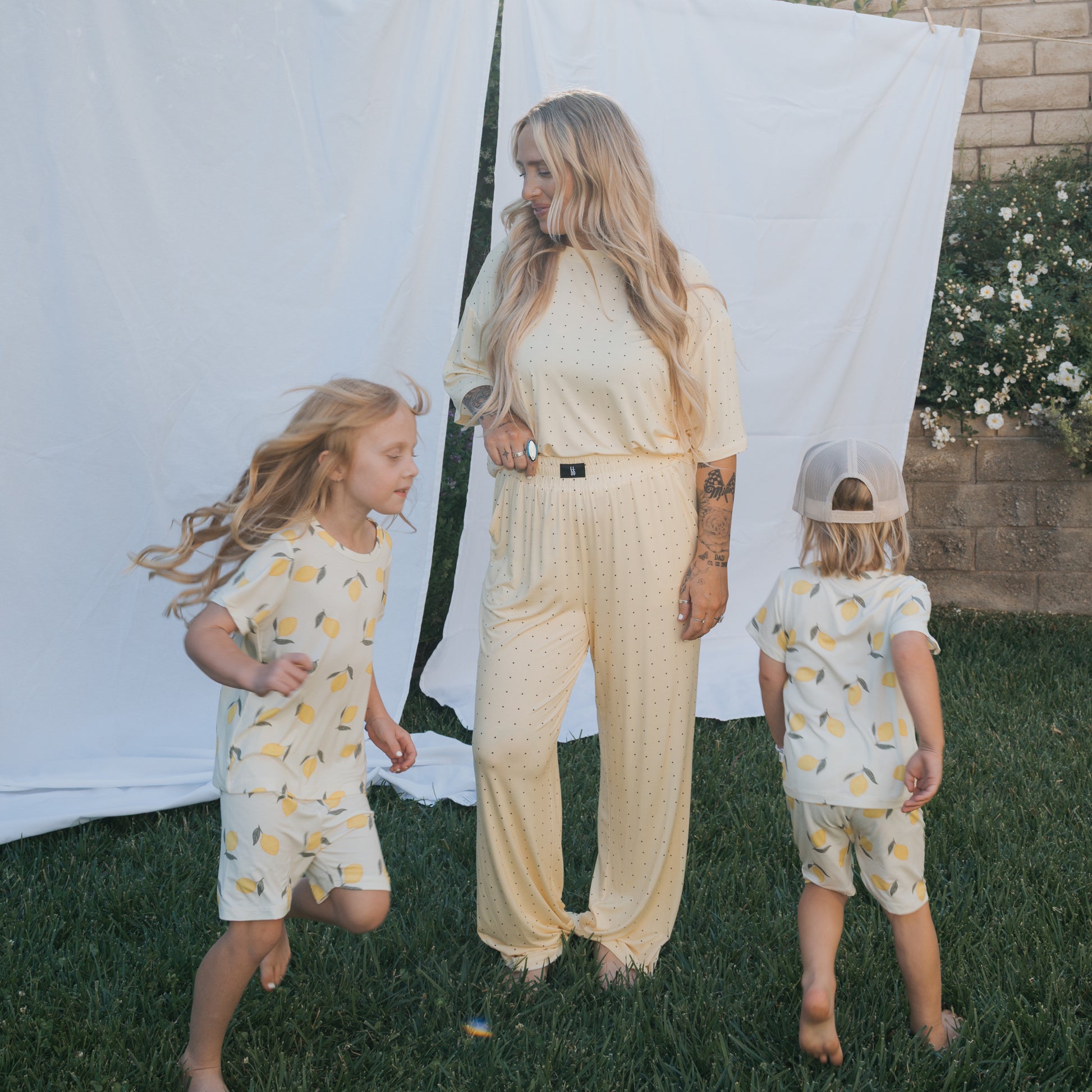 A woman in a Women's Boxy Tee Lounge Set by forever french baby stands on grass, watching two children in matching bamboo pajamas play with a white sheet and blooming flowers in the background.