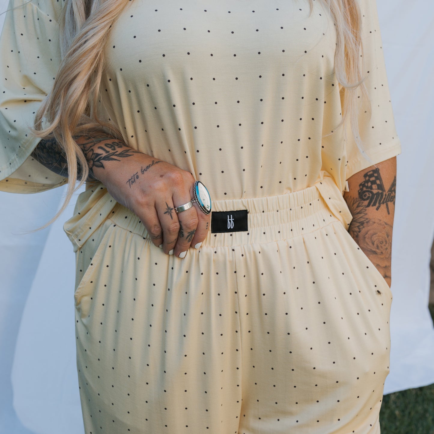 A person with long blonde hair wears the forever french baby Women's Boxy Tee Lounge Set | Veda in light yellow polka dots, one hand in their pocket, displaying arm tattoos and a large ring.