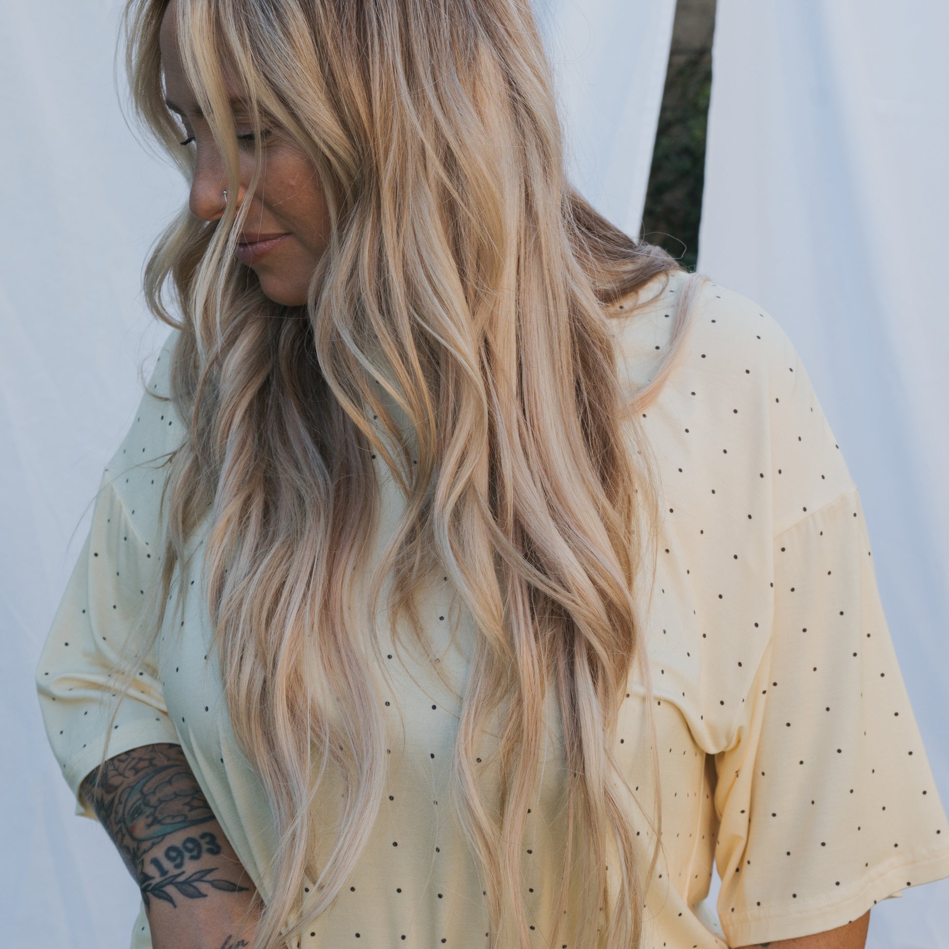 A woman with long, wavy blonde hair wearing the forever french baby Women's Boxy Tee Lounge Set in light yellow with black dots looks down, showing a tattoo on her arm. White fabric drapes in the background.