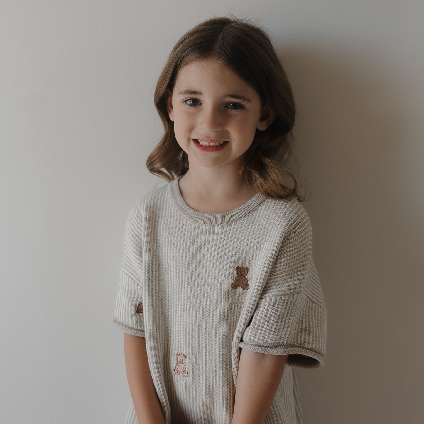 A young girl with brown hair smiles against a plain white wall, wearing the forever french baby Knit Short Set | Teddy—featuring cream-colored, striped soft knit fabric with small teddy bear designs.