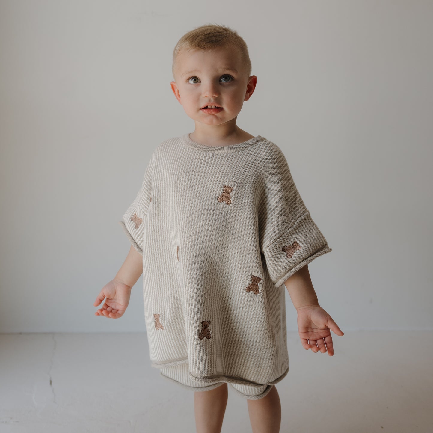 A young child stands indoors wearing the forever french baby Knit Short Set | Teddy: an oversized cream cotton top and drawstring shorts, both with teddy bear designs, made from soft knit. The well-lit background is plain.