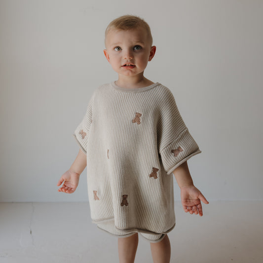 A young child stands indoors wearing the forever french baby Knit Short Set | Teddy: an oversized cream cotton top and drawstring shorts, both with teddy bear designs, made from soft knit. The well-lit background is plain.