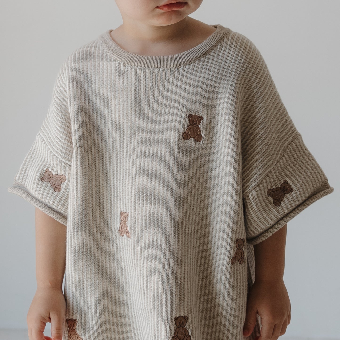 A young child in the forever french baby Knit Short Set | Teddy—a loose, beige ribbed knit outfit with small brown teddy bears—stands against a plain light background, their face partially out of frame.