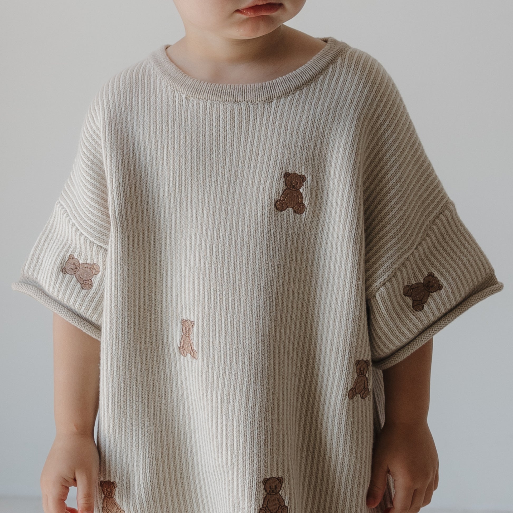 A young child in the forever french baby Knit Short Set | Teddy—a loose, beige ribbed knit outfit with small brown teddy bears—stands against a plain light background, their face partially out of frame.