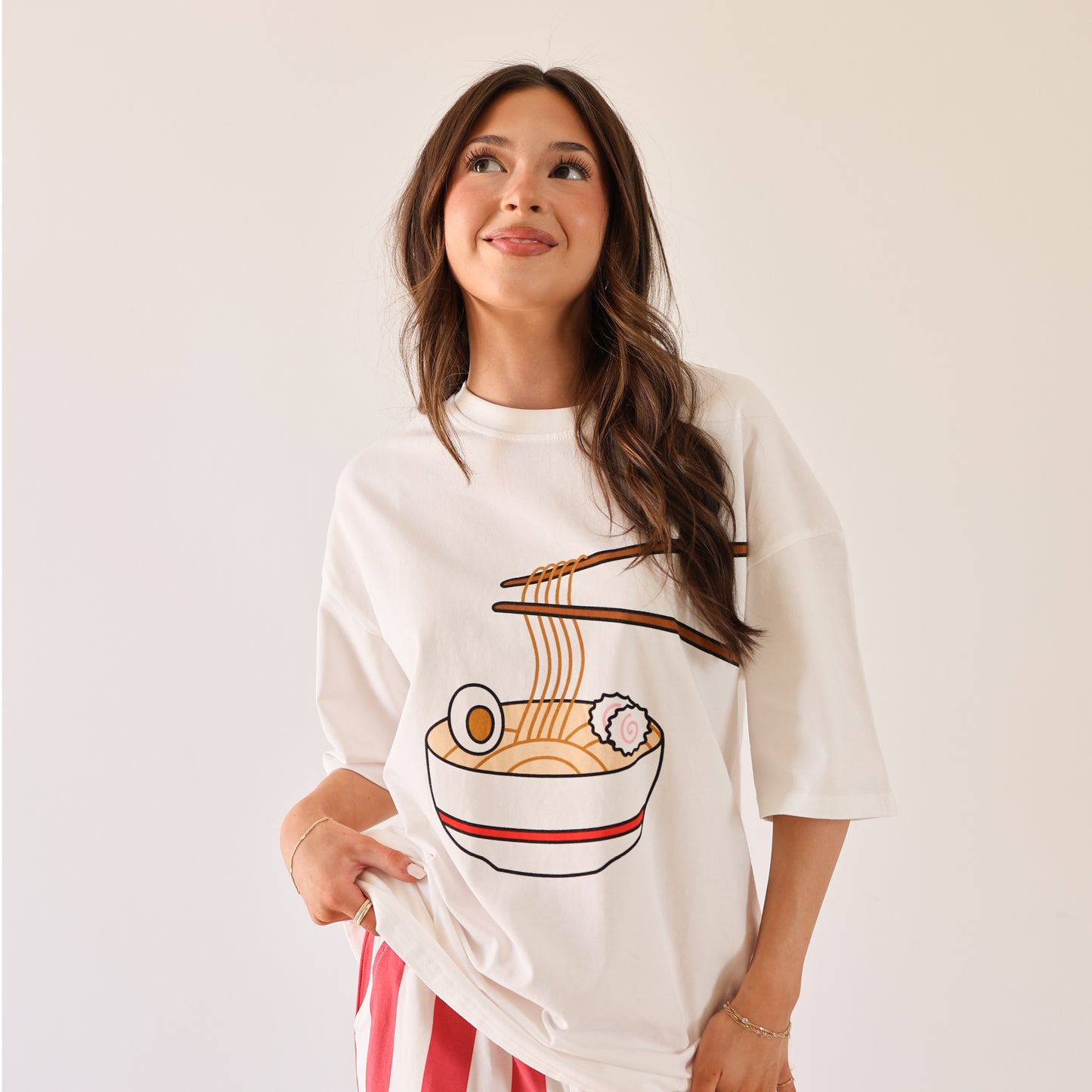 A woman with long brown hair smiles slightly in a plain setting, wearing the forever french baby Adult Cotton Tee | Ramen, which is super soft and features a graphic of chopsticks holding ramen noodles above a bowl.