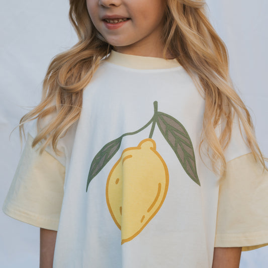 A young girl with long blonde hair smiles slightly, wearing the forever french baby Child Cotton Tee | Lemon—crafted from 100% cotton with a bold yellow lemon and green leaves—showcasing stylish summer kidswear against a plain, light background.