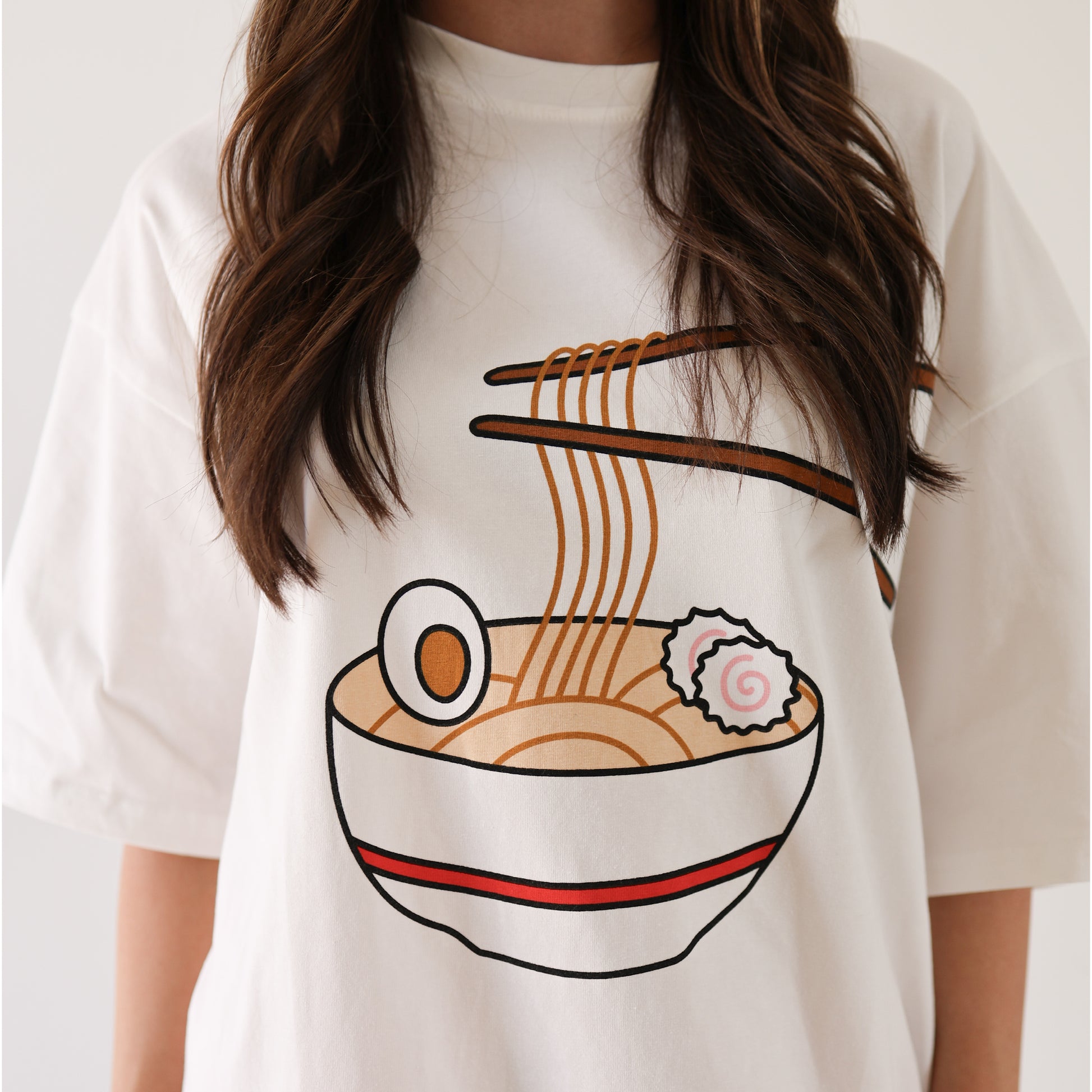 A person (face hidden) wears the forever french baby Adult Cotton Tee | Ramen, a super soft white shirt featuring a graphic of ramen noodles, boiled egg, and narutomaki being lifted by chopsticks.