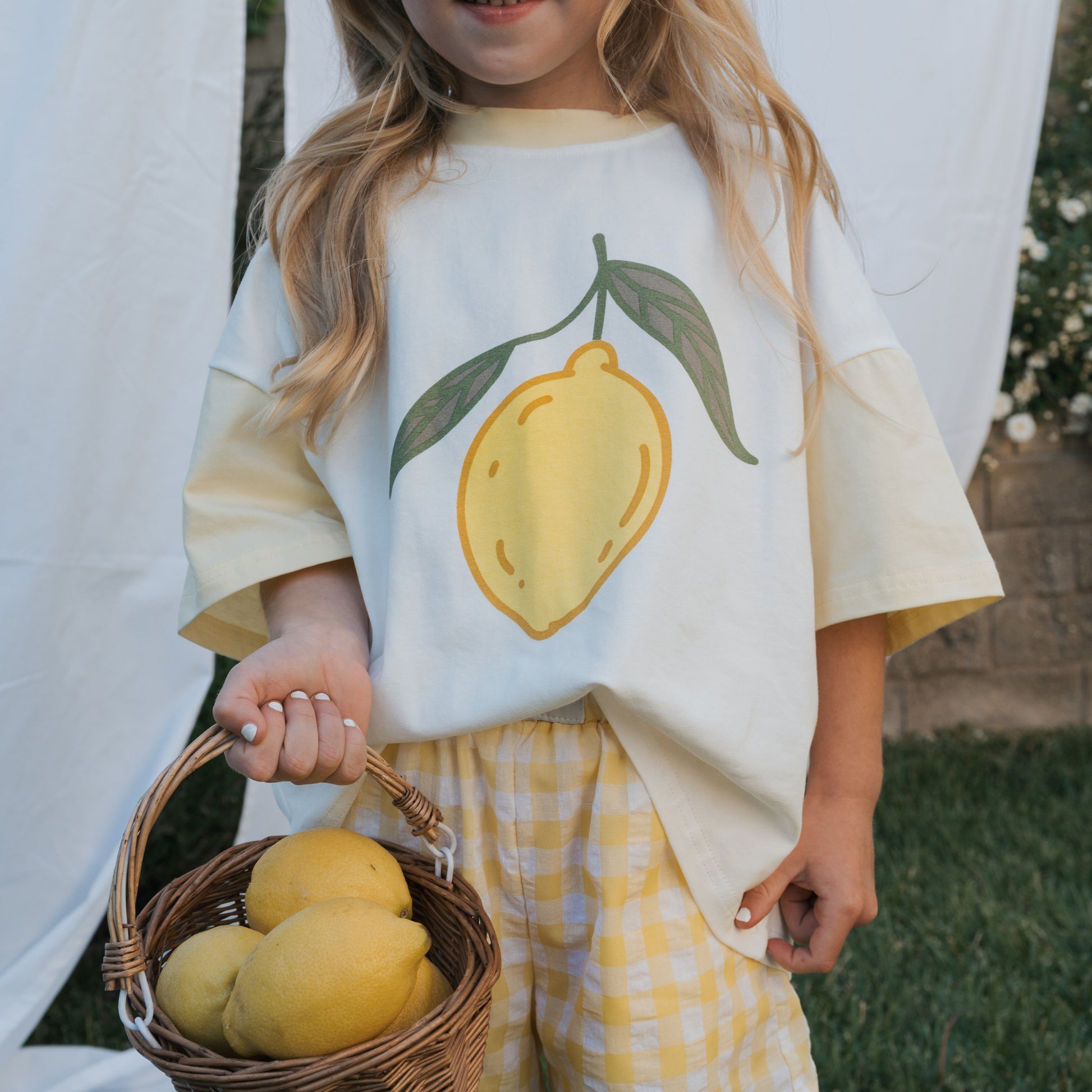 A young girl wears the forever french baby Child Cotton Tee | Lemon with yellow checkered shorts, holding a basket of lemons outdoors among white fabric and greenery—embodying the charm of summer kids’ fashion.