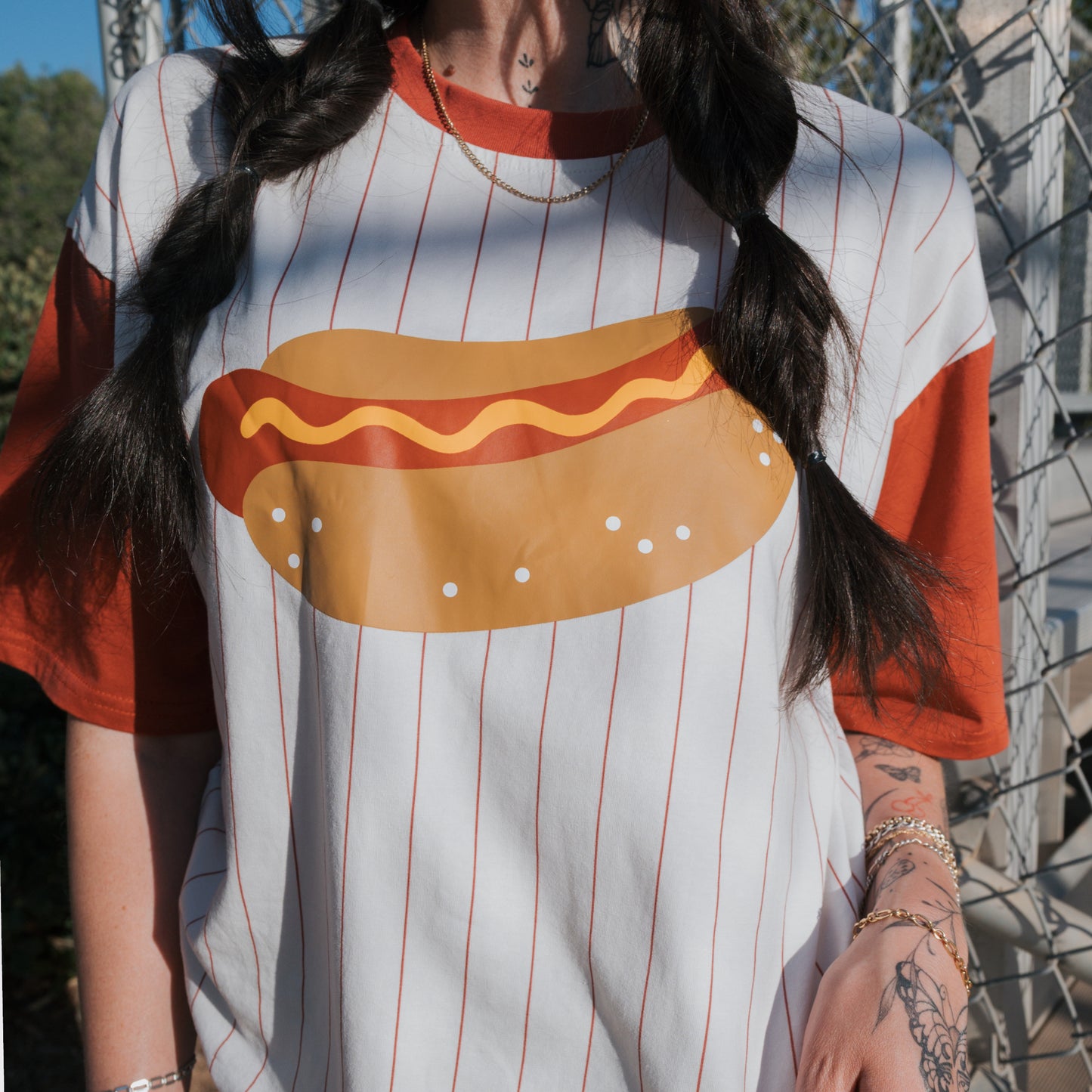 A person with braided hair and left-arm tattoos stands outdoors by a metal fence, wearing the forever french baby Adult Cotton Tee | Hot Dogs—a super soft white shirt with red pinstripes and a large hot dog graphic with mustard.