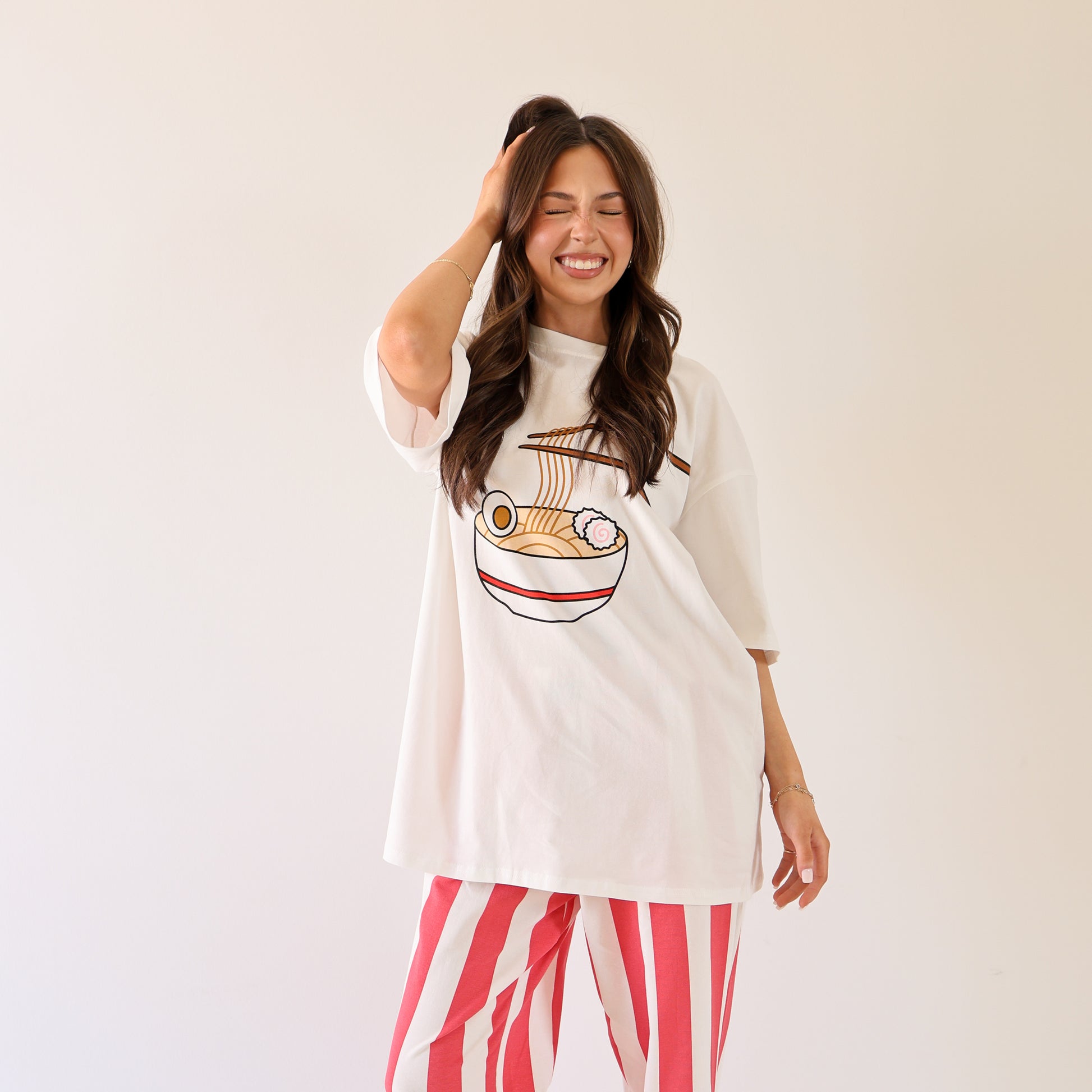 A woman with long brown hair smiles with her eyes closed, wearing the forever french baby Adult Cotton Tee | Ramen, which features a ramen bowl illustration and an oversized fit, paired with red-and-white striped pants against a plain background.