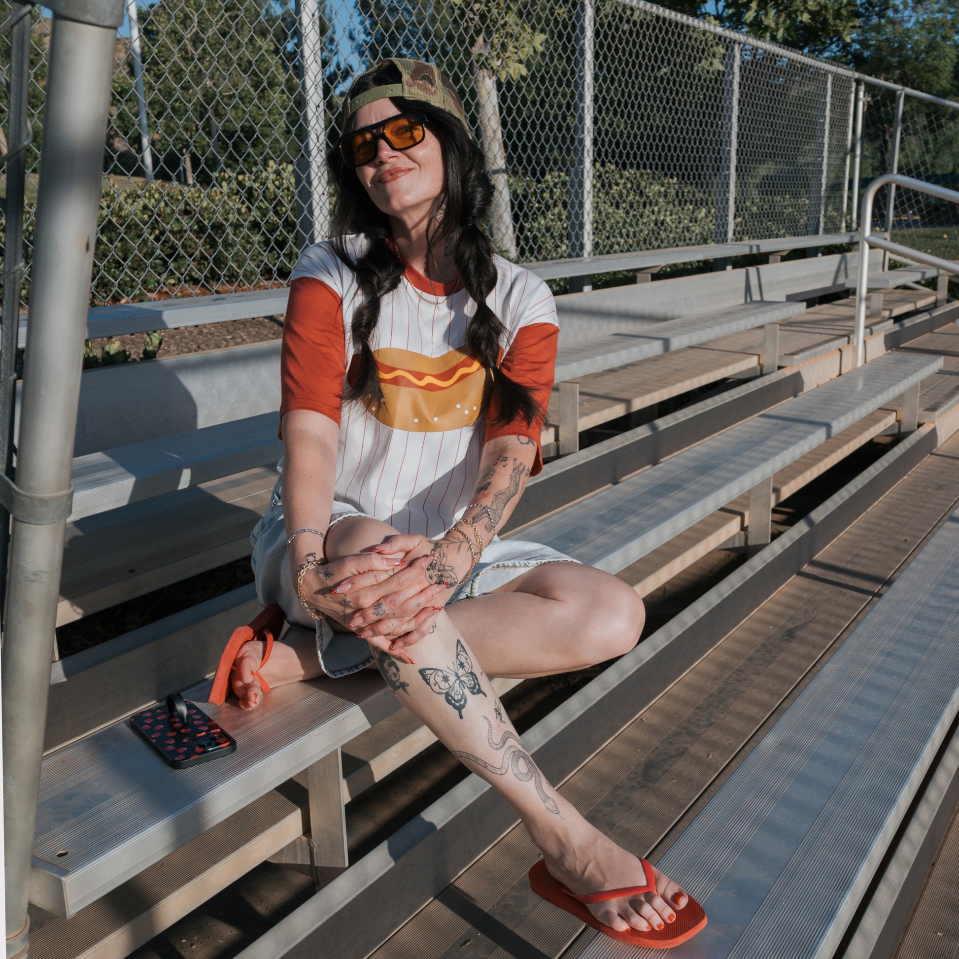 A woman wears the forever french baby Adult Cotton Tee | Hot Dogs, plus sunglasses and a cap, sitting on bleachers. Her arms and legs are tattooed; red flip-flops and a phone are by her side on a sunny day.