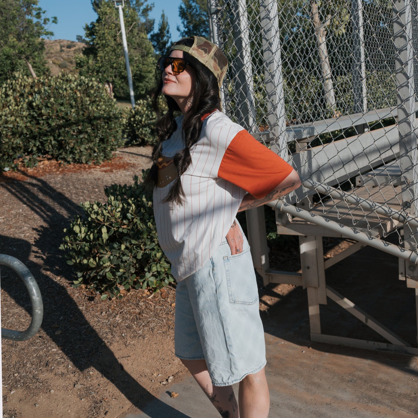 A person with long dark hair, sunglasses, and a camo cap stands by metal bleachers outdoors, wearing the forever french baby Adult Cotton Tee | Hot Dogs with orange sleeves and light denim shorts, hands behind their back.