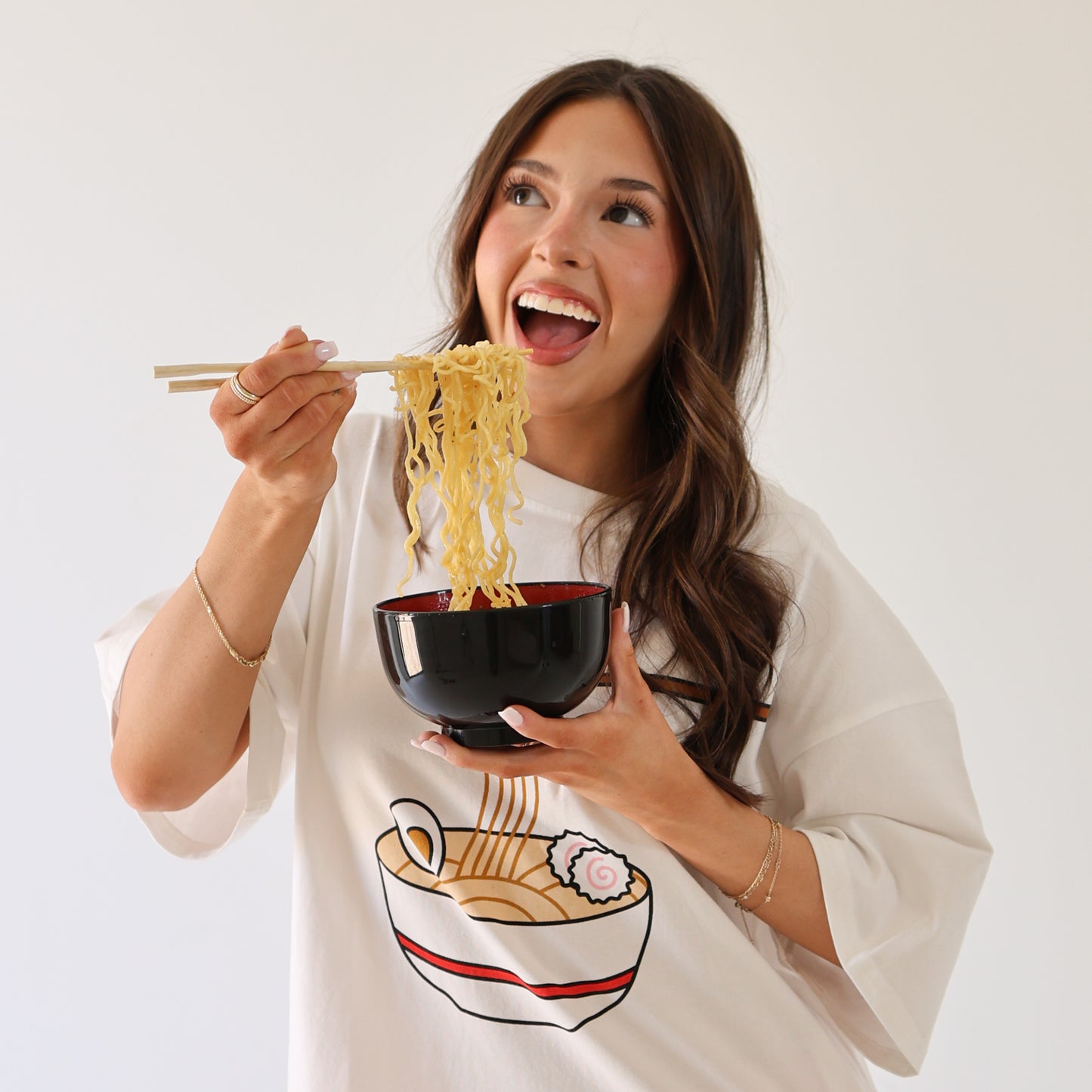 A smiling woman with long brown hair lifts noodles from a black bowl while wearing the forever french baby Adult Cotton Tee | Ramen, which features a cartoon ramen bowl graphic, against a plain white background.