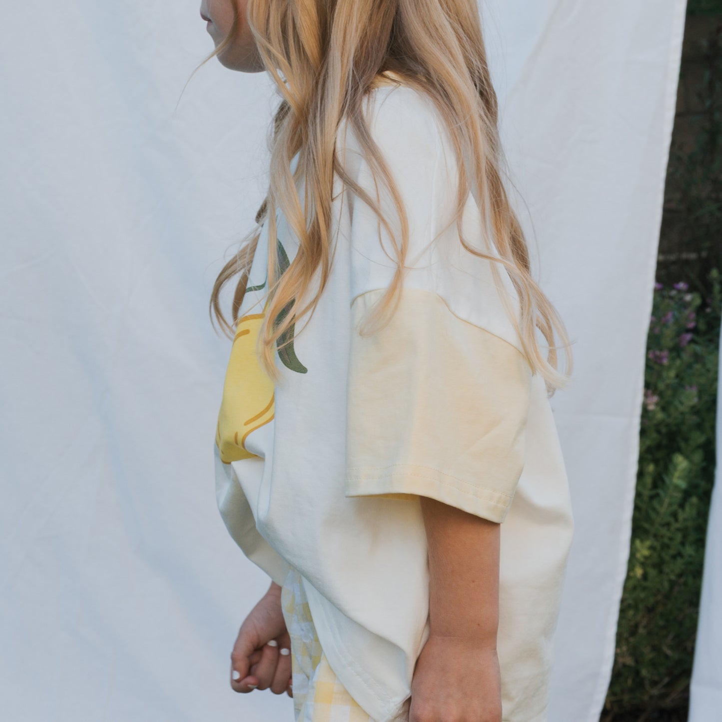 A child with long blonde hair stands sideways outdoors before a white backdrop, wearing the forever french baby Child Cotton Tee | Lemon—made from 100% cotton and perfect for summer kids clothing.