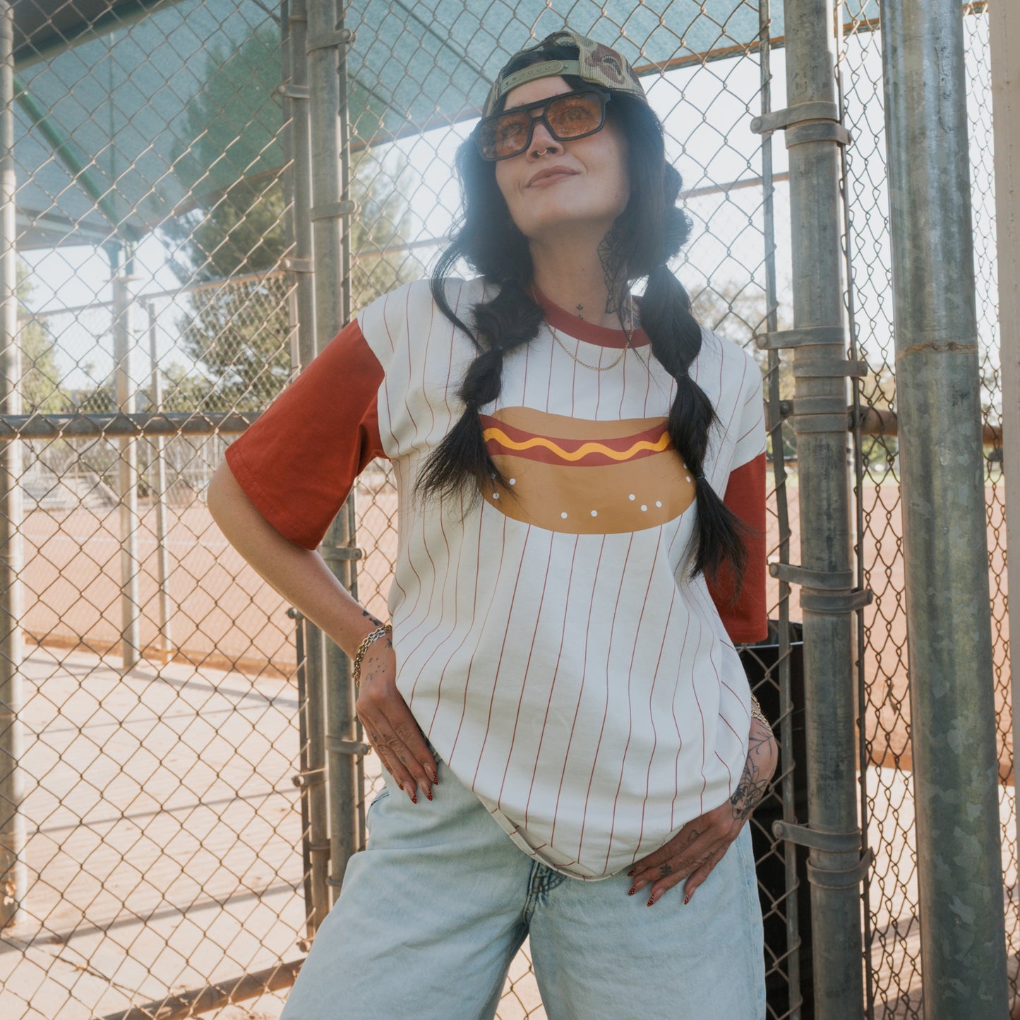 Wearing sunglasses, a backwards cap, and a forever french baby Adult Cotton Tee | Hot Dogs, someone poses confidently by a chain-link fence at an outdoor sports field.