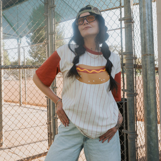 Wearing sunglasses, a backwards cap, and a forever french baby Adult Cotton Tee | Hot Dogs, someone poses confidently by a chain-link fence at an outdoor sports field.