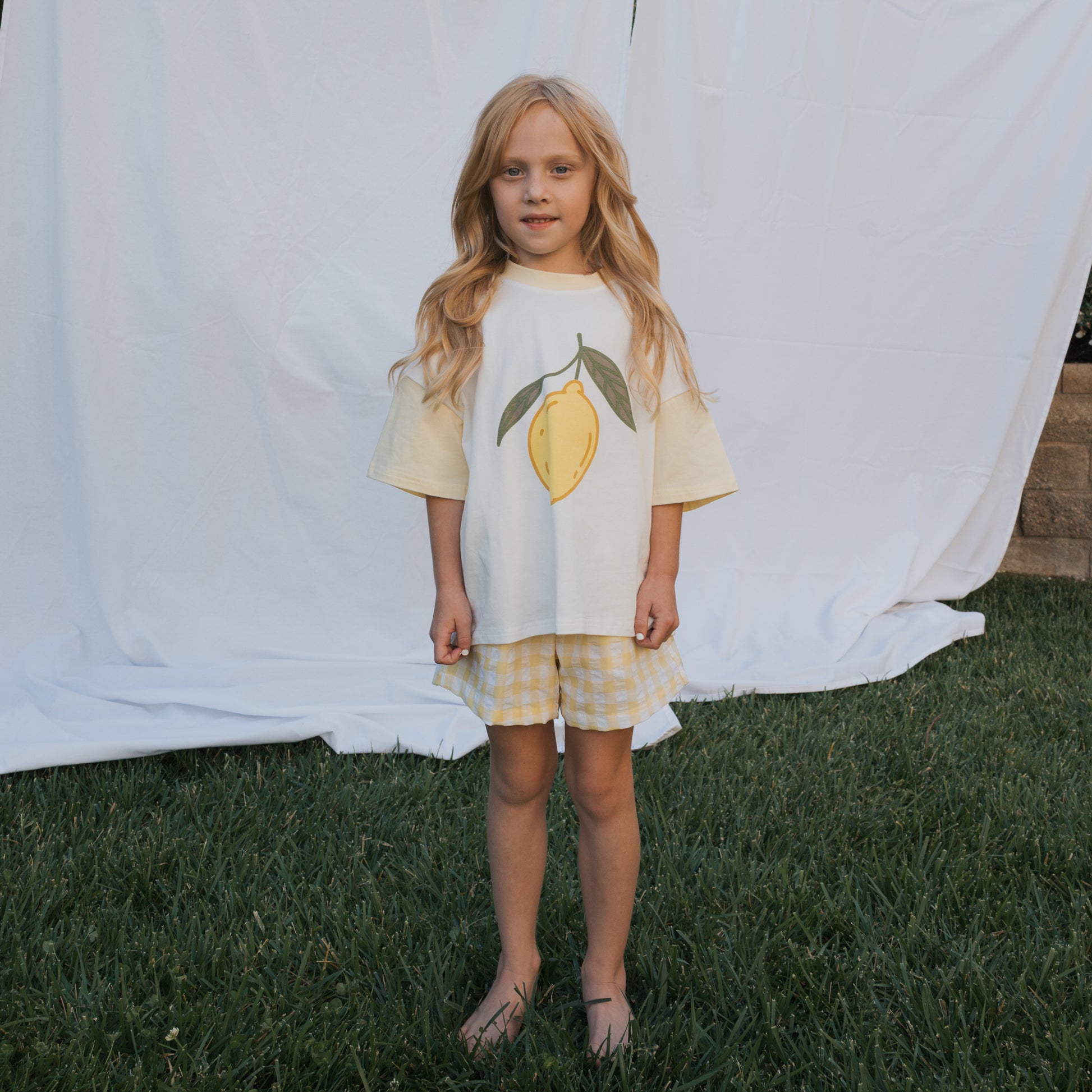 A young girl with long blonde hair stands barefoot on grass, wearing the forever french baby Child Cotton Tee | Lemon and yellow plaid shorts—perfect summer kids’ clothing. A white sheet is draped behind her as a backdrop.