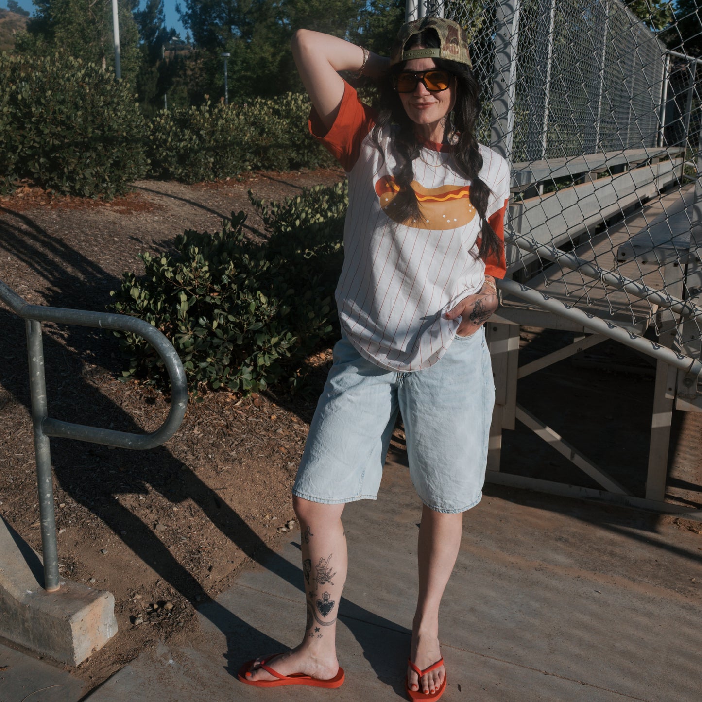 A person wearing sunglasses, a camouflage cap, the forever french baby Adult Cotton Tee | Hot Dogs, denim shorts, and red flip-flops stands outdoors by metal bleachers, posing with one hand behind their head.