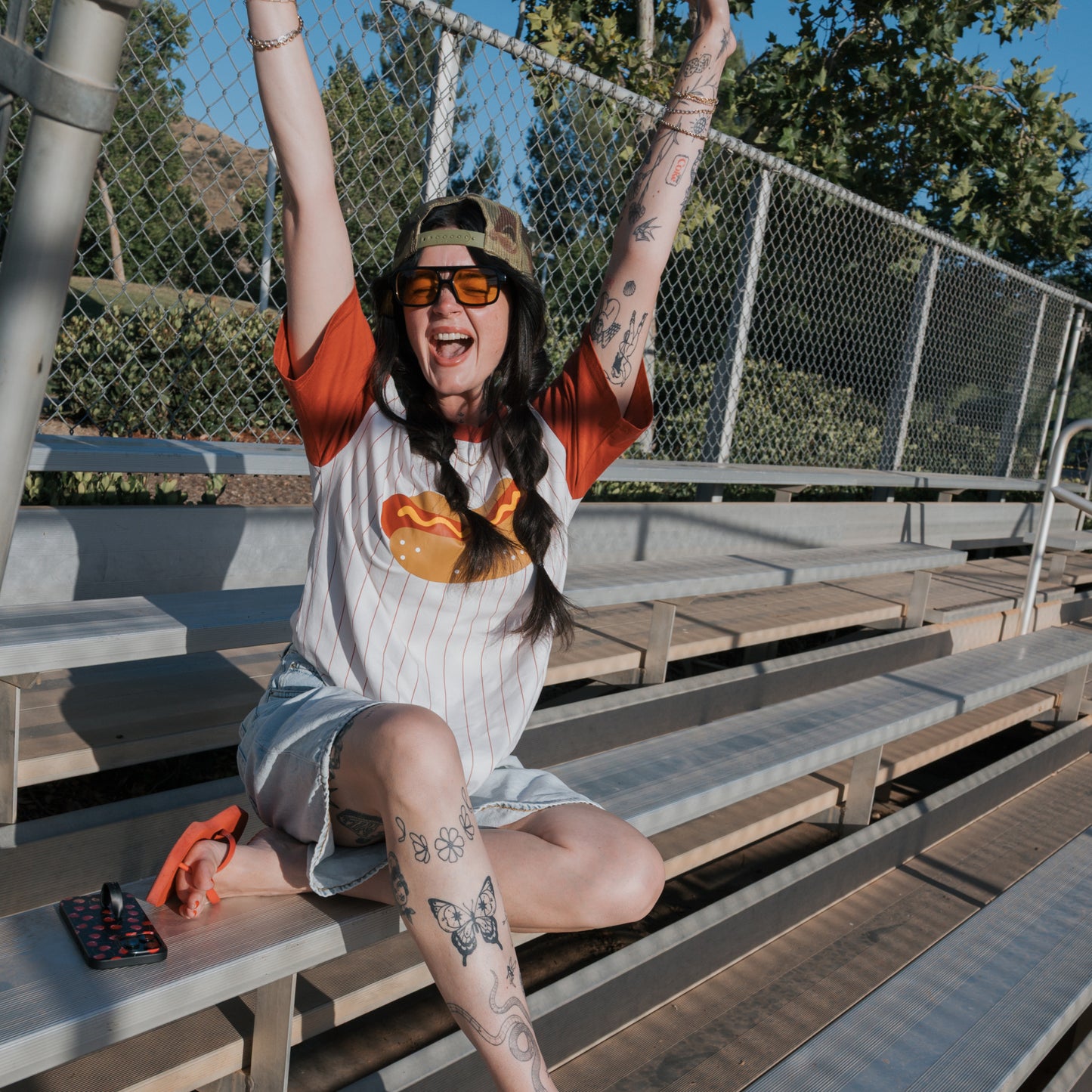 Wearing the forever french baby Adult Cotton Tee | Hot Dogs, a tattooed person in sunglasses and a cap smiles on bleachers with arms raised. Sunlight and greenery brighten the background.