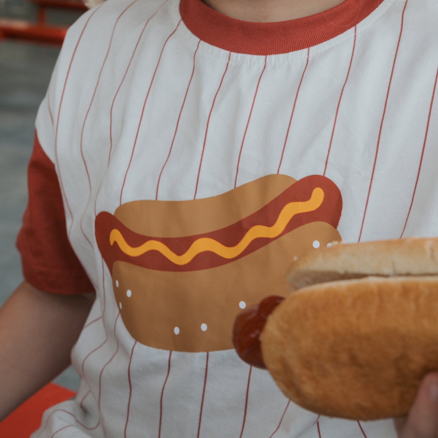 A person wearing the "Child Cotton Tee | Hot Dogs" by forever french baby, featuring a cartoon hot dog and red raglan sleeves, casually holds a real hot dog in their hand.