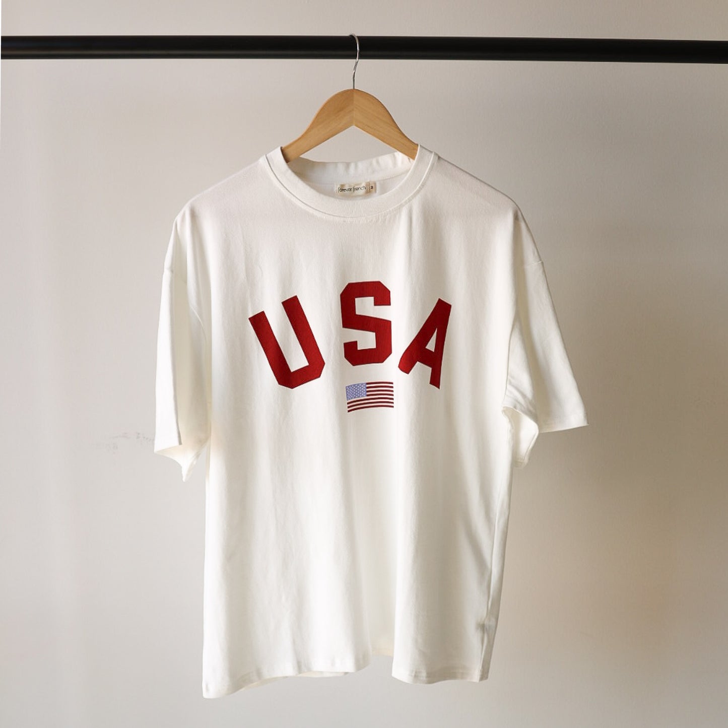 The forever french baby Adult Cotton Tee | USA is a 100% cotton unisex t-shirt on a wooden hanger, featuring bold red "USA" lettering with a small American flag graphic below.